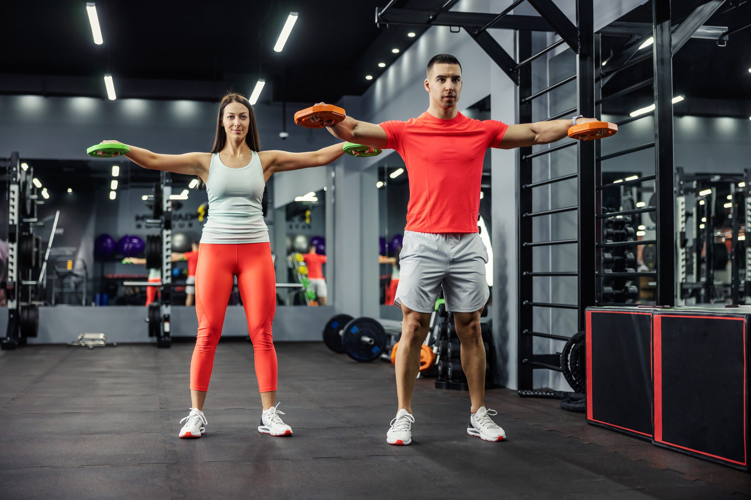 A muscular man and a beautiful young woman doing some arm and shoulder exercises together in the indoor gym at night time. Couple goals, fitness goals